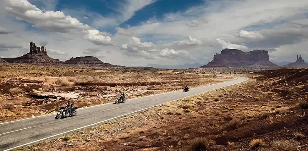 Motociclistas conduciendo Harley Davidson en carretera en desierto con montañas pedrosas de fondo